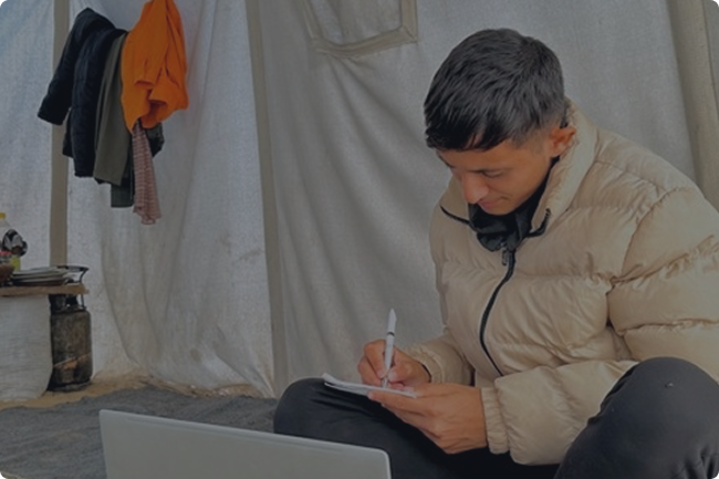 A student writing in a notebook beside a laptop inside a tent